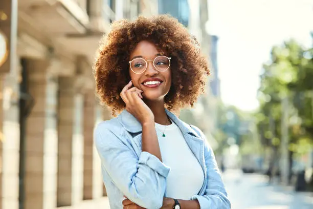 Smiling young woman talking on smart phone. Beautiful female is wearing eyeglasses on sidewalk. She is having frizzy hair.
