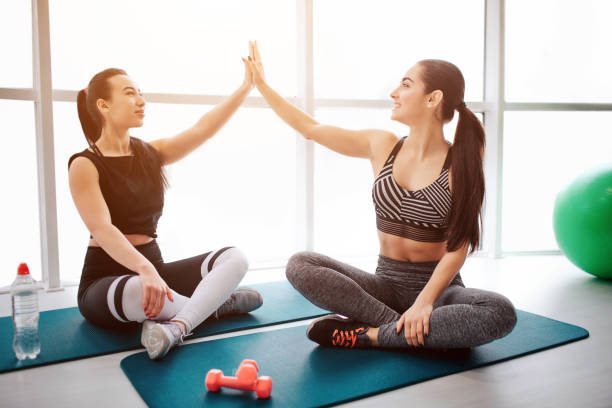 Beautiful and well-built young women sit on matrass in lotus pose. They give high-five to each other. Models sit in fitness room. Beautiful and well-built young women sit on matrass in lotus pose. They give high-five to each other. Models sit in fitness room matrass stock pictures, royalty-free photos & images