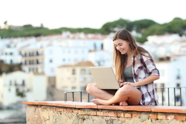 istock Happy teen using a laptop sitting on a ledge on a town 1091381010