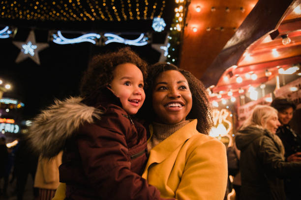 Mother and Daughter at Christmas Markets A close-up front-view shot of a woman holding her young daughter, they are both looking at bright​ Christmas lights at the Christmas markets on a cold night, the young girl is in awe. christmas market photos stock pictures, royalty-free photos & images