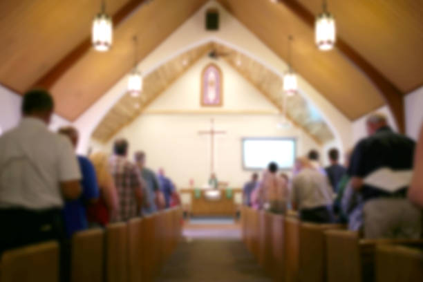 Blurred Photo of the Iterior of a Church Sanctuary with Congregation, Pastor, and a Large Cross Visible A blurred photo of the inside of a church sanctuary that is filled with people in the pews, and the pastor stands under a large cross at the altar. clergy stock pictures, royalty-free photos & images