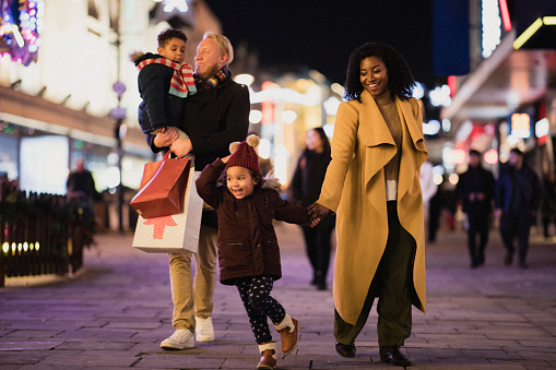 A front-view shot of a multi-ethnic family with two children walking down a city street on a cold December night.