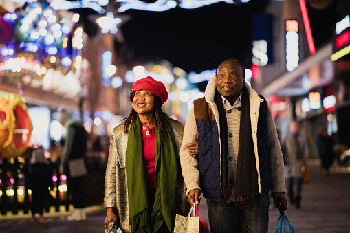 A front-view shot of a senior couple walking down a city street, they are smiling and wearing warm clothing on the cold December​ night.