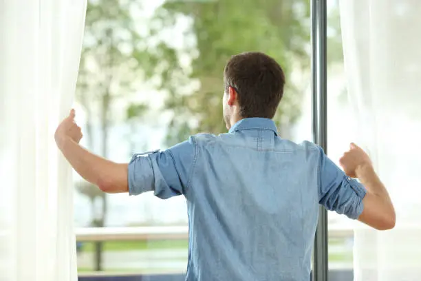 Photo of Back view of man opening curtains and looking away