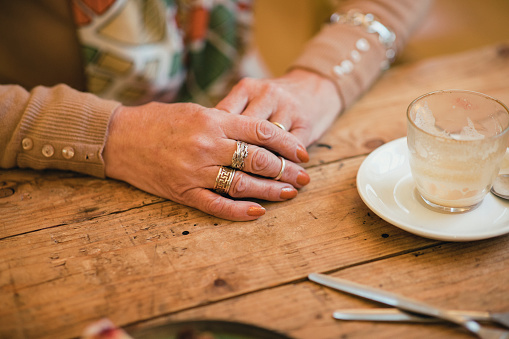 Close-up of an unrecognisable senior woman hands clasped on a table. She has multiple rings on different fingers.