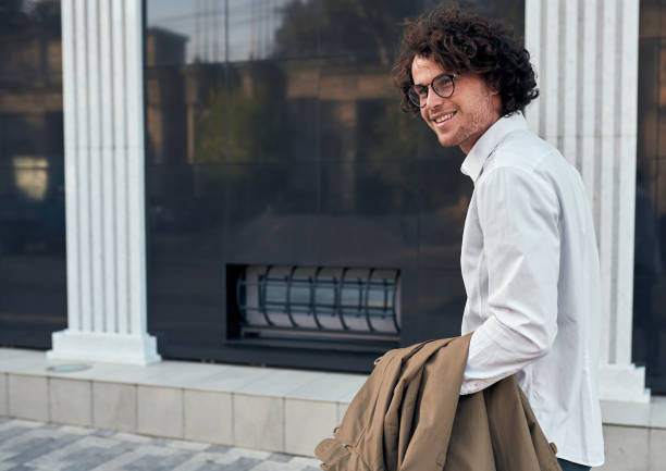 tiro de vista lateral do jovem empresário positivo com óculos posando ao ar livre. masculino universitário na rua. cara esperto em camisa casual veste branco e óculos com cabelo encaracolado, andando na rua - smart casual side view one person success - fotografias e filmes do acervo