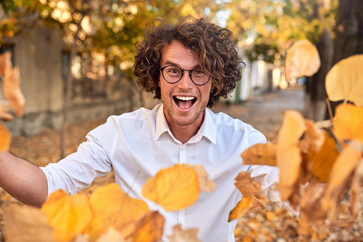 Horizontal autumn shot of happy handsome winner young man with glasses playing with leaves outdoors. Successful male student enjoy triumph. Guy in white shirt, spectacles with curly hair on street