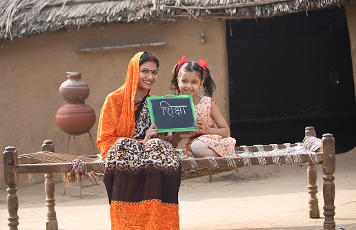 Happy Indian mother with her daughter holding slate while sitting on traditional bed in village