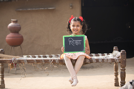 Little Indian girl holding slate while sitting on traditional bed in village