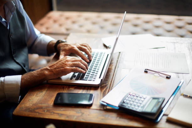 senior zakenman te typen op een laptop in een naaiatelier ruimte - financiën en economie stockfoto's en -beelden