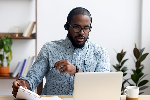 Black young man sitting at table wearing headphones learn foreign language improves knowledge looking at pc screen listening audio lesson holding pen and notepad makes some notes. E-learning concept
