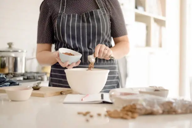 Shot of woman wearing apron adding cocoa powder in bowl at kitchen counter.