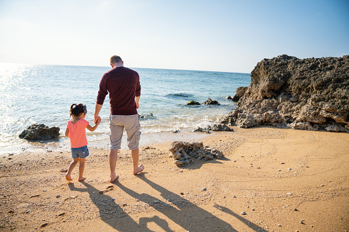 Father and young daughter walking along and exploring a beach while holding hands during a vacation