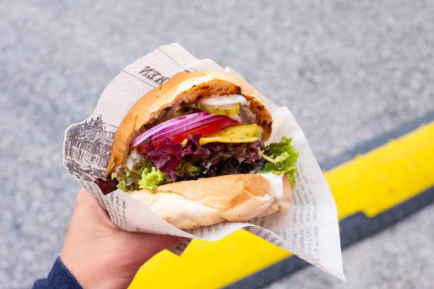 hand eines jungen mannes, der einen biologisch vegane burger mit seitan patty in seiner hand hält, während ein suppen-festival - fleischersatz stock-fotos und bilder