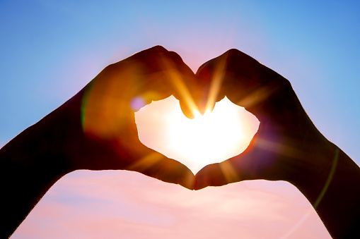 A DSLR photo of hands making a heart shape with the sun with beautiful sunbeams inside. Blue sky background. Space for copy.