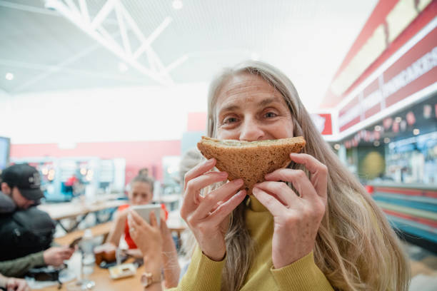 One mature woman is posing for the camera in the airport terminal, holding half of a vegan sandwich to her face like a mouth.