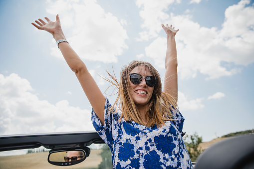 Young woman is sitting in a convertible car with her arms in the air while on holiday.