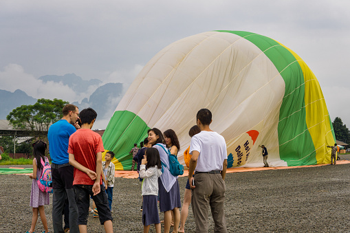 Vang Vieng, Laos - October 3, 2018:  A hot air balloon is landing.  Bright green and yellow in colour, it has flown over the mountains in the distance.  Here we see several tourists waiting for a trip in the balloon.  Hot air balloon trips are a popular tourist activity in Vang Vieng.