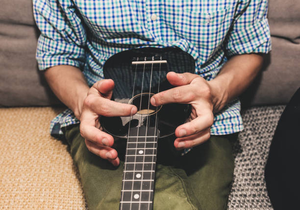 Man in checkered shirt at home is playing the ukulele guitar. Guys hands are taking the chord on strings. Music making lifestyle concept. Free time hobby for everyone. Funny guitar photo. hombre de camisa a cuadros en la casa es tocar la guitarra ukelele. manos de chicos están tomando el acorde en las cuerdas. concepto de estilo de vida que hace de la música. afición de tiempo libre para todos. foto de guitarra divertido.