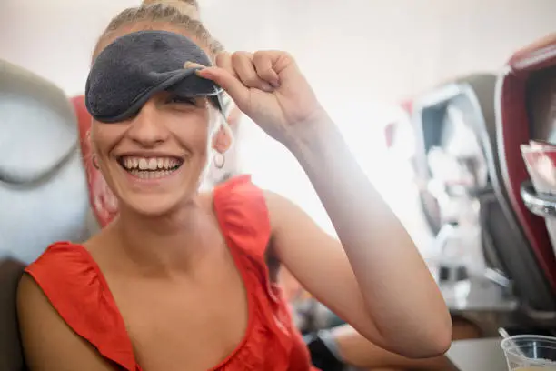One young woman has woken up from a nap on a flight and is peeking out through her eye mask looking at the camera with a toothy smile.