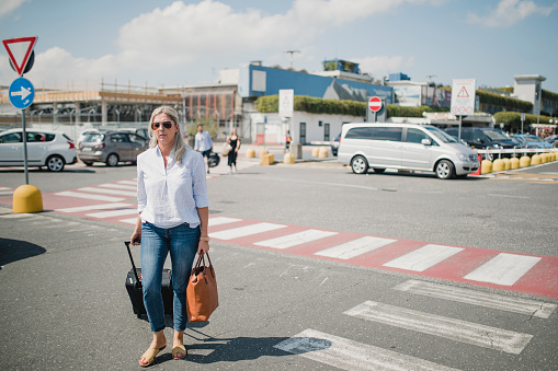 One mature woman is walking unhappily through the parking lot of the airport with her baggage.