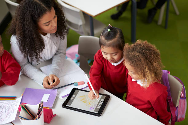 Elevated view of two girls using a tablet computer sitting at table in an infant school class a with female teacher helping them Elevated view of two girls using a tablet computer sitting at table in an infant school class a with female teacher helping them teacher classroom child education stock pictures, royalty-free photos & images