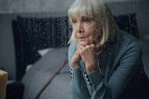 zdenerwowany starsza kobieta siedzi i podpierając podbródek z rękami w domu przez okno z kroplami deszczu - propping zdjęcia i obrazy z banku zdjęć