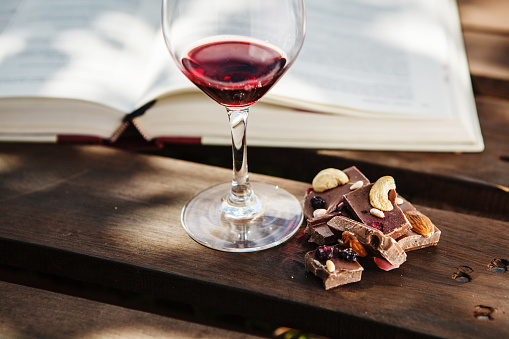 Glass with red wine and pieces of chocolate with nuts and raisins stands on wooden bar on background of open book. Alcoholic drink in glassware with snack