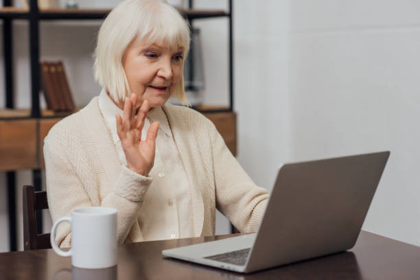 istock senior woman sitting at computer desk and waving while having video call at home 1091720142