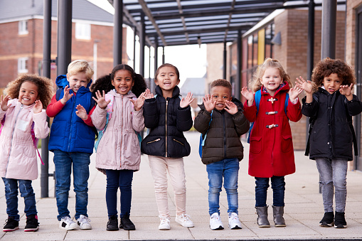 Un grupo de jóvenes escuela multiétnica de sonreír a los niños vistiendo abrigos y transporte de mochilas de pie en una fila en el pasillo fuera de su escuela de niño saludando a la cámara, largo, vista frontal photo