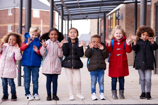 笑顔若い多民族学校グループ子供着てコートやカメラ、完全な長さ、正面に手を振って自分の幼児学校の外の通路の行に立っているランドセル キャリング - coat ストックフォトと画像
