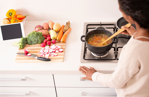 High angle, over the shoulder view of a woman stirring the boiling bouillon, using a wooden spoon. The woman cooking vegetable soup in a stock pot on a gas stove top, using a digital tablet to follow the recipe online. The tablet computer learning on a bowl with bell peppers. Sliced radish, broccoli, raw potato, carrot, ginger, and zucchini are on cutting board. A healthy meal, lifestyle, and culinary concepts.