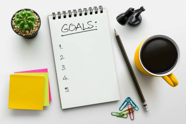istock Top view notebook with goals list. Notepad on office table with a cup of coffee, plant, stationery and office supplies. Top view desk, flat lay white background concept. 1091752236