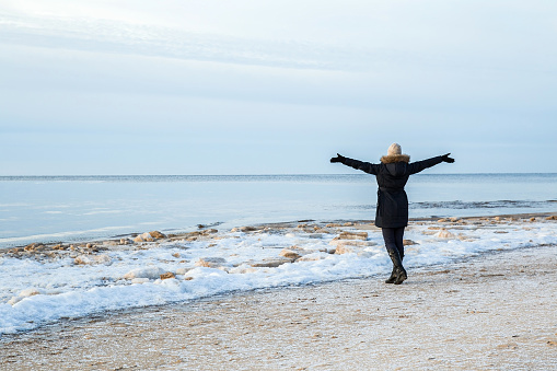 Young adult woman with outstretched arms standing on sea beach in winter day. Peaceful atmosphere. Back view.