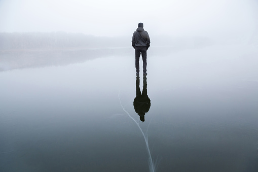 Young adult man standing alone on cracked dark ice surface. Mist over frozen lake in winter. Foggy air. Early chilly morning. Peaceful atmosphere in nature. Back view.