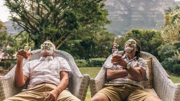 Elderly men with facial mask and cucumber slices on their face at spa. Retired friends enjoying facial spa treatment with juice glasses in hand.