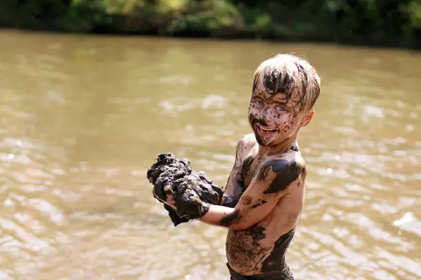 A happy young boy child is covered in mud as he laughs, swims, and plays outside in the river on a summer day.