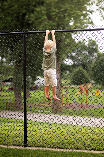 A Little boy child was trying to climb a tall chain link fence to secape to a playground, but is stuck hanging by his hands at the top.