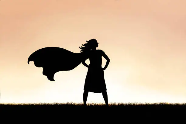 The silhouette of a strong, beautiful caped super hero woman stands isolated against a sunset in the sky background.