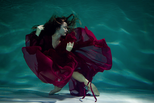 Dancing woman under the water in a pool in a red dress.