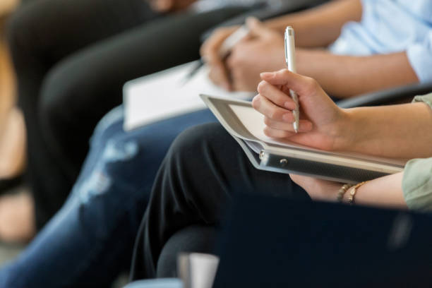 Unrecognizable business person takes notes during training class In this closeup, no one is recognizable as a business person sits in a row of coworkers during a training class.  She holds a pen ready to take notes.  There is also a binder in her lap. presentation folder stock pictures, royalty-free photos & images