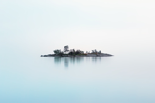 Remote rocky island with coniferous trees as if hanging in the misty air over the milky surface of the lake in a summer night. Atmospheric cool northern background with copy space. White Nights season, Lake Onega, Karelia, Russia.