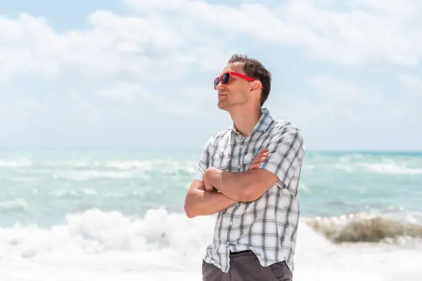 Young man hipster millennial arms crossed on beach during sunny day with red sunglasses in Miami, Florida with ocean in background