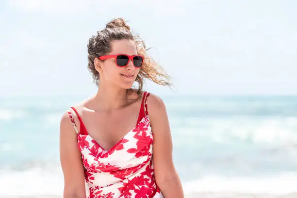 Young beautiful woman in red and white floral summer dress sitting on beach during sunny day with sunglasses in Miami Florida, looking to side smiling happy