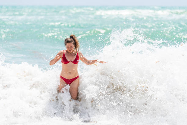 istock Young woman standing in red swimsuit bikini bathing suit scared surprised by splash crashing wave in Hollywood, Miami Beach Florida green water drops 1091910496