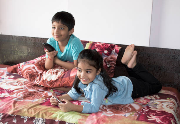 Girl using mobile phone while brother is watching tv on bed Sibling relaxing on bed with TV remote control and mobile phone indian boy barefoot stock pictures, royalty-free photos & images