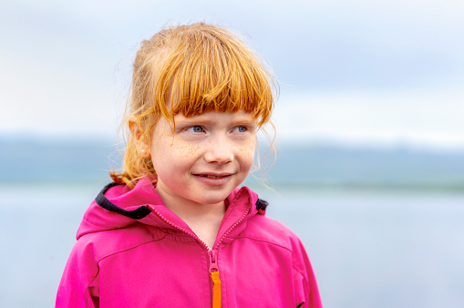 Close-up of smiling cute girl looking away. Redhead female is contemplating against lake. He is wearing pink jacket.