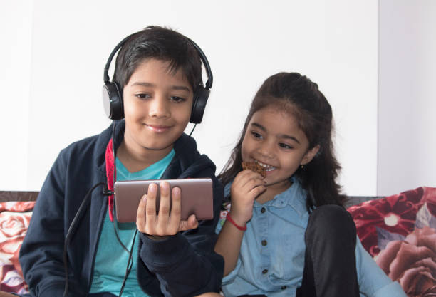 crianças usando o telefone celular e fones de ouvido enquanto come biscoito em casa - indian girls audio - fotografias e filmes do acervo