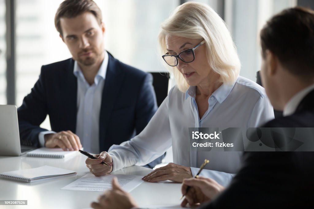 Concentrated aged businesswoman checking agreement before signing Businessmen sitting at desk headed by middle aged serious concentrated female in eyeglasses checking agreement document before signing it. Financial director ready affirm official paper with signature Contract Stock Photo