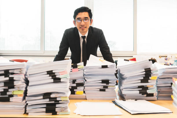 Businessman in suit standing in office with pile of documents on foreground. Businessman in suit standing in office with pile of documents on foreground paper clip office supply stack heap stock pictures, royalty-free photos & images